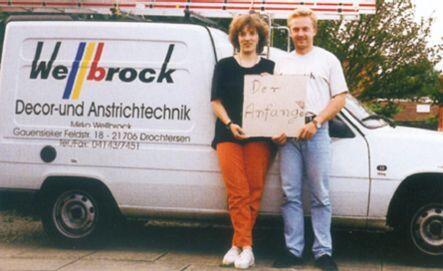 Frau und Mann stehen vor einem Auto mit der Aufschrift "Wellbrock Decor- und Anstrichtechnik" und halten ein Schild in der Hand auf dem "Der Anfang" steht